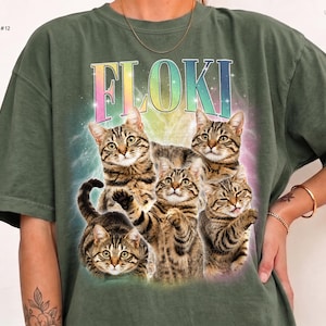 May include: A moss green short-sleeve t-shirt featuring a graphic print of four brown tabby cats. Above the cats, the text FLOKI is printed in a rainbow gradient font. The background of the graphic includes a soft, glowing, multi-colored light effect.