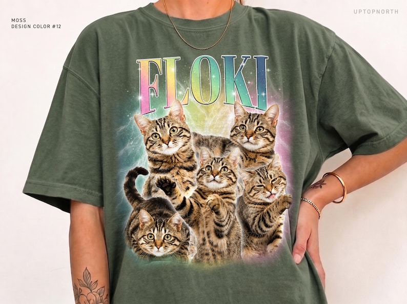 May include: A moss green short-sleeve t-shirt featuring a graphic print of four brown tabby cats. Above the cats, the text FLOKI is printed in a rainbow gradient font. The background of the graphic includes a soft, glowing, multi-colored light effect.
