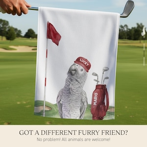 Op de afbeelding: Een wit golfhanddoekje met wafelstructuur voorzien van een bedrukte illustratie van een grijze papegaai met een rode pet met het woord LUCKY, naast een rode golfbag met de naam MARIO en een vlaggetje op een green.