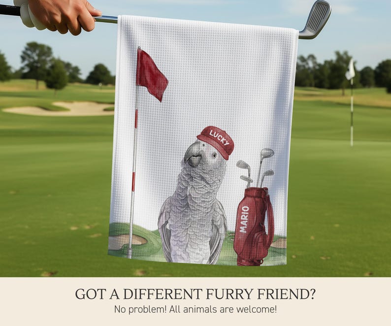 Op de afbeelding: Een wit golfhanddoekje met wafelstructuur voorzien van een bedrukte illustratie van een grijze papegaai met een rode pet met het woord LUCKY, naast een rode golfbag met de naam MARIO en een vlaggetje op een green.