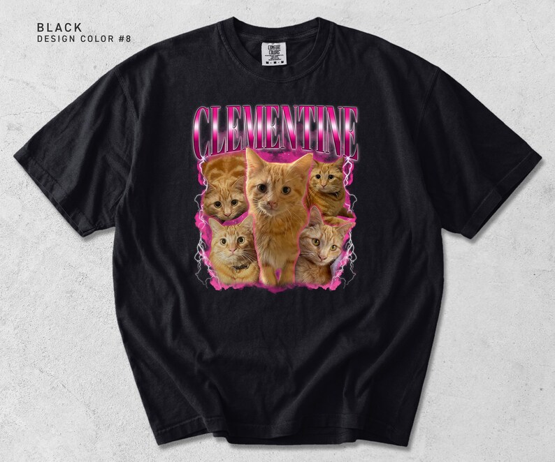 May include: A black short-sleeve t-shirt featuring a graphic print of five orange cats. The text CLEMENTINE is printed in a metallic pink font above the cats, which are surrounded by a pink lightning bolt design. The shirt has a crew neckline.