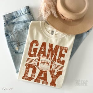 May include: An ivory t-shirt with the words "GAME DAY" in distressed orange lettering and a football graphic. The shirt is laid on light-wash denim jeans, a tan hat, and a necklace. The shirt is from Comfort Colors.