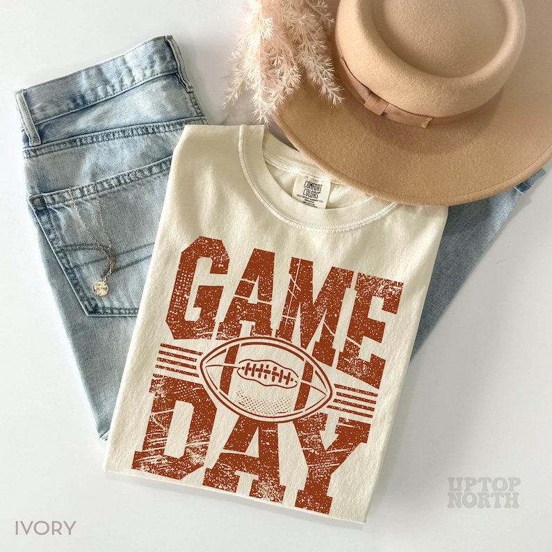 May include: An ivory t-shirt with the words "GAME DAY" in distressed orange lettering and a football graphic. The shirt is laid on light-wash denim jeans, a tan hat, and a necklace. The shirt is from Comfort Colors.