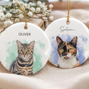 May include: Two round white ceramic ornaments with gold hanging strings. One features a brown tabby cat illustration with the name OLIVER, the other a calico cat illustration with the name Salem. Both have watercolor-style backgrounds.