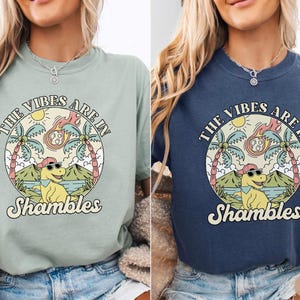 May include: Two t-shirts, one in bay green and the other in denim blue, feature a circular graphic. The graphic includes a dinosaur wearing sunglasses and a hat, palm trees, and the text "THE VIBES ARE IN Shambles".