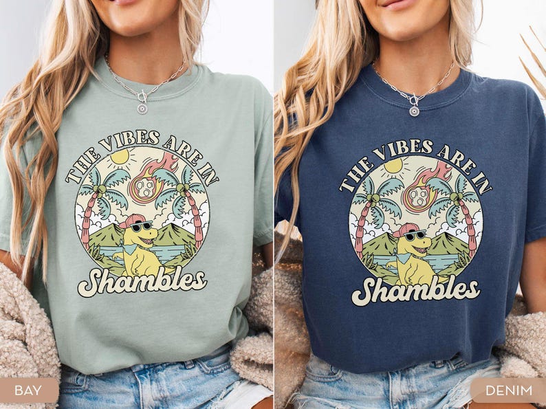 May include: Two t-shirts, one in bay green and the other in denim blue, feature a circular graphic. The graphic includes a dinosaur wearing sunglasses and a hat, palm trees, and the text "THE VIBES ARE IN Shambles".