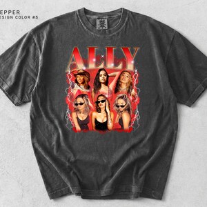 K&ouml;nnte beinhalten: Ein dunkelgraues T-Shirt mit einem Retro-Grafikdesign. Das Wort "ALLY" ist in Rot gehalten, mit Bildern von sechs Frauen. Das T-Shirt hat einen Rundhalsausschnitt und kurze &Auml;rmel. Das Design ist von Vintage inspiriert.