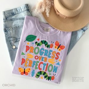 Op de afbeelding: Een lichtpaars t-shirt met de tekst "PROGRESS OVER PERFECTION" in kleurrijke letters. Het shirt heeft illustraties van rupsen, vlinders en een blad. Het shirt wordt gecombineerd met een spijkerbroek, een ketting en een beige hoed.