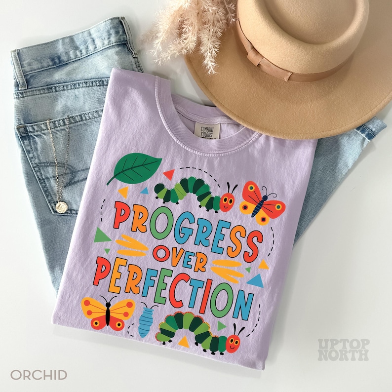 Op de afbeelding: Een lichtpaars t-shirt met de tekst "PROGRESS OVER PERFECTION" in kleurrijke letters. Het shirt heeft illustraties van rupsen, vlinders en een blad. Het shirt wordt gecombineerd met een spijkerbroek, een ketting en een beige hoed.