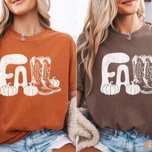 May include: Two t-shirts, one yam-colored and one espresso-colored, each featuring a white graphic. The graphic includes the word "FALL", cowboy boots, and pumpkins. The shirts are short-sleeved and have a vintage, distressed look.