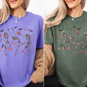 K&ouml;nnte beinhalten: Zwei kurz&auml;rmelige T-Shirts mit Rundhalsausschnitt, eines in Violett und eines in Moosgr&uuml;n, jeweils mit einem Grafikdruck von drei tanzenden Skeletten zwischen orangefarbenen Herbstbl&auml;ttern, schwarzen Flederm&auml;usen und kleinen K&uuml;rbiseimern.