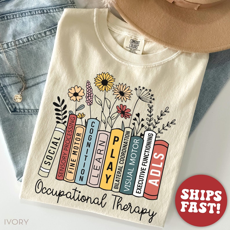 May include: A beige t-shirt with a graphic of a stack of colorful books with text that reads "SOCIAL", "SENSORY PROCESSING", "FINE MOTOR", "COGNITION", "LEARN", "PLAY", "BILATERAL COORDINATION", "VISUAL MOTOR", "EXECUTIVE FUNCTIONING", and "ADLS". The books are surrounded by flowers and the text "Occupational Therapy" is below the books.