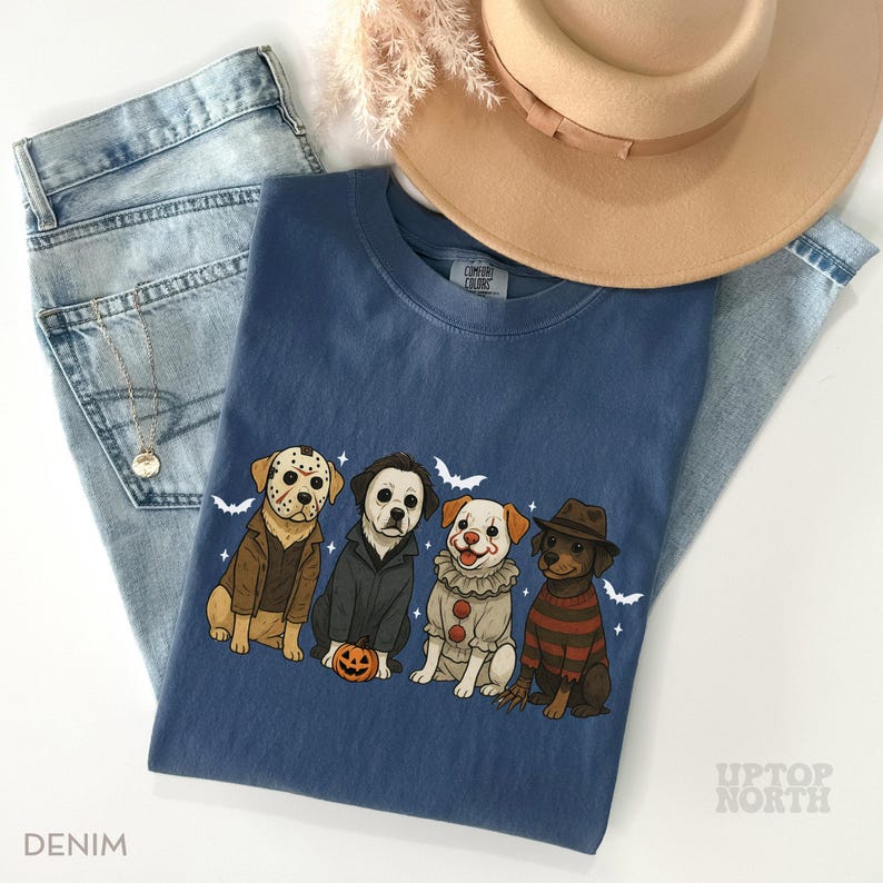 May include: A denim blue Comfort Colors t-shirt featuring a Halloween-themed graphic of four dogs dressed as horror movie characters. The graphic includes a dog in a Jason Voorhees mask, Michael Myers, a clown, and Freddy Krueger. The shirt is paired with light wash jeans and a tan hat.