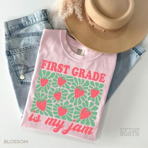 May include: A light pink t-shirt with the text "FIRST GRADE is my jam" in red and green. The shirt features a strawberry design. The shirt is laid flat with a pair of blue jeans and a tan hat.
