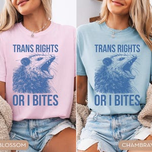 Puede incluir: Dos camisetas, una rosa claro y otra azul claro, cada una con el texto "TRANS RIGHTS" sobre una ilustraci&oacute;n azul de una zarig&uuml;eya y las palabras "OR I BITES" debajo.