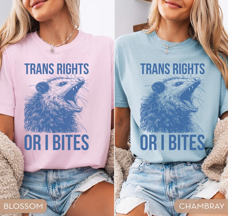 Puede incluir: Dos camisetas, una rosa claro y otra azul claro, cada una con el texto "TRANS RIGHTS" sobre una ilustraci&oacute;n azul de una zarig&uuml;eya y las palabras "OR I BITES" debajo.