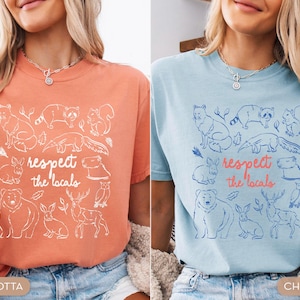 K&ouml;nnte beinhalten: Terracotta- und chambrayblaue T-Shirts mit einer Grafik verschiedener Tiere und dem Text "respect the locals". Das Design umfasst einen Fuchs, einen Waschb&auml;ren, ein Eichh&ouml;rnchen, ein Kaninchen, einen B&auml;ren, ein Reh und eine Eule. Die Shirts sind kurz&auml;rmelig.