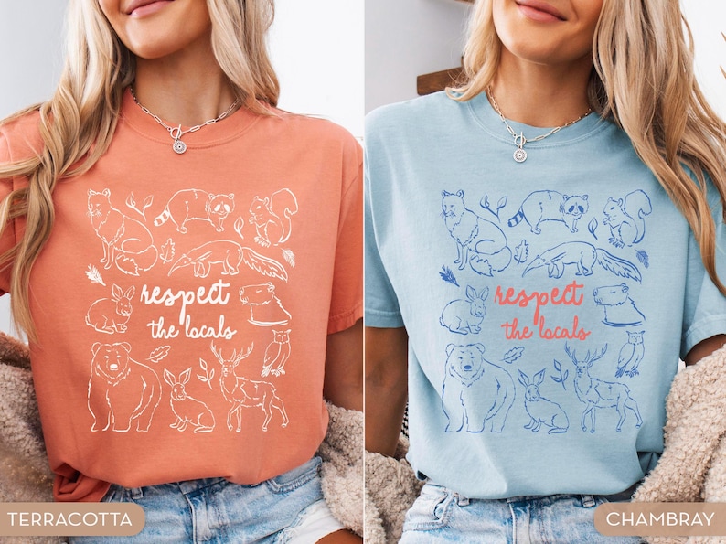 K&ouml;nnte beinhalten: Terracotta- und chambrayblaue T-Shirts mit einer Grafik verschiedener Tiere und dem Text "respect the locals". Das Design umfasst einen Fuchs, einen Waschb&auml;ren, ein Eichh&ouml;rnchen, ein Kaninchen, einen B&auml;ren, ein Reh und eine Eule. Die Shirts sind kurz&auml;rmelig.