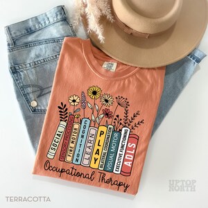 May include: Terracotta t-shirt with a graphic of a stack of books with the words "Occupational Therapy" and the words "Social, Sensory Processing, Fine Motor, Cognition, Learn, Play, Bilateral Coordination, Visual Motor, ADLS, Executive Functioning" on the books. The books are surrounded by flowers.
