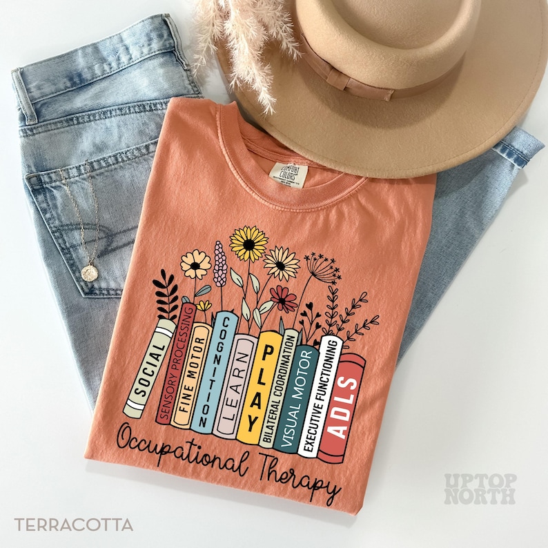 May include: Terracotta t-shirt with a graphic of a stack of books with the words "Occupational Therapy" and the words "Social, Sensory Processing, Fine Motor, Cognition, Learn, Play, Bilateral Coordination, Visual Motor, ADLS, Executive Functioning" on the books. The books are surrounded by flowers.