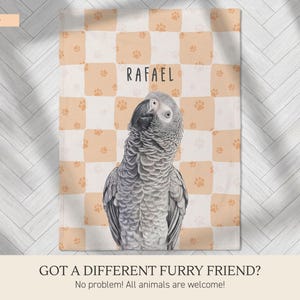 May include: A rectangular throw blanket featuring a light peach and white checkered pattern with small orange paw prints. The center displays a realistic illustration of a grey African Grey parrot and the name RAFAEL in black text.