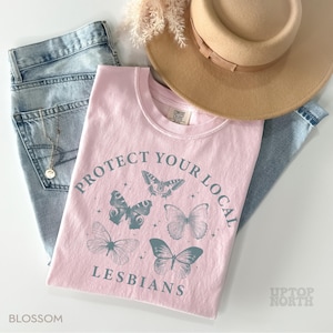 May include: A light pink t-shirt with the text "PROTECT YOUR LOCAL LESBIANS" and butterfly graphics. The shirt is paired with light blue jeans and a tan hat. The word "BLOSSOM" is printed at the bottom left corner.
