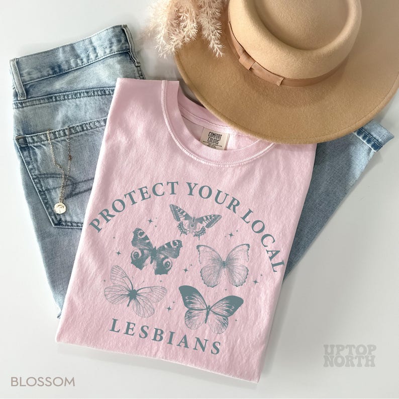May include: A light pink t-shirt with the text "PROTECT YOUR LOCAL LESBIANS" and butterfly graphics. The shirt is paired with light blue jeans and a tan hat. The word "BLOSSOM" is printed at the bottom left corner.