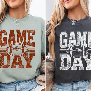 May include: Two t-shirts, one light green and one dark grey, each with a distressed "GAME DAY" design. The design includes a football graphic. The shirts are casual wear.