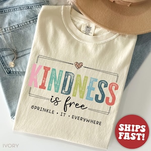 Pode incluir: Camiseta de cor marfim com o texto "Kindness is free. Sprinkle it everywhere" em cores do arco-&iacute;ris. Um pequeno cora&ccedil;&atilde;o de estampa de leopardo est&aacute; acima do texto.