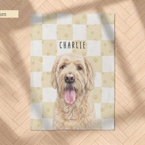 May include: A cream and white checkered blanket featuring a repeating pattern of small paw prints and a central illustration of a golden-colored dog with its tongue out. The name CHARLIE is printed in black capital letters above the dog.