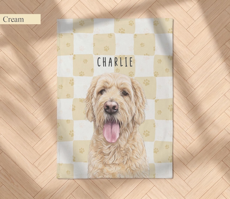 May include: A cream and white checkered blanket featuring a repeating pattern of small paw prints and a central illustration of a golden-colored dog with its tongue out. The name CHARLIE is printed in black capital letters above the dog.