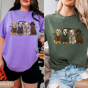 May include: Two t-shirts, one violet and one moss green, each featuring a graphic of four dogs dressed as horror movie characters. The dogs are wearing costumes and are sitting in a row with bats above them.