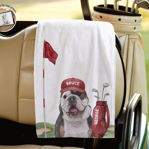 Op de afbeelding: Een witte golfhanddoek met een grafische illustratie van een buldog die een rode pet draagt met de naam BRUCE erop gedrukt. Naast de hond staat een rode golf-tas met het label GEORGE en een rode golfvlag aan een vlaggenmast.