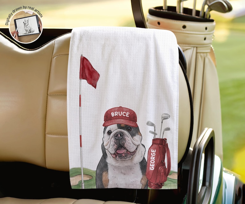Op de afbeelding: Een witte golfhanddoek met een grafische illustratie van een buldog die een rode pet draagt met de naam BRUCE erop gedrukt. Naast de hond staat een rode golf-tas met het label GEORGE en een rode golfvlag aan een vlaggenmast.