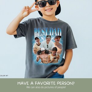 May include: A charcoal gray short-sleeve t-shirt featuring a graphic print with the text BEST DAD in blue and white. The design includes a collage of five photographs of a man in various settings, surrounded by a glowing blue light effect.