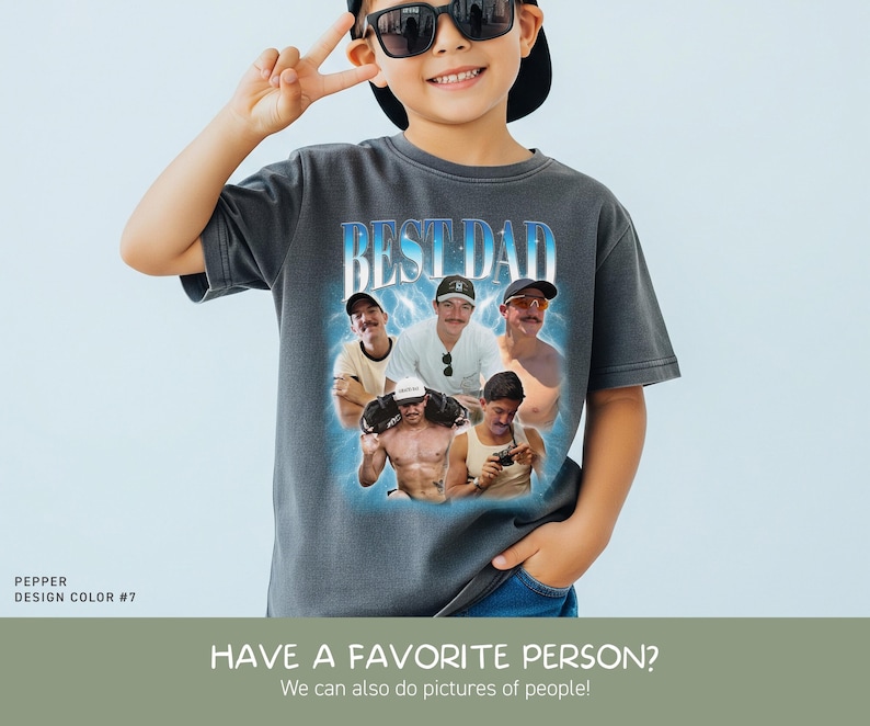 May include: A charcoal gray short-sleeve t-shirt featuring a graphic print with the text BEST DAD in blue and white. The design includes a collage of five photographs of a man in various settings, surrounded by a glowing blue light effect.