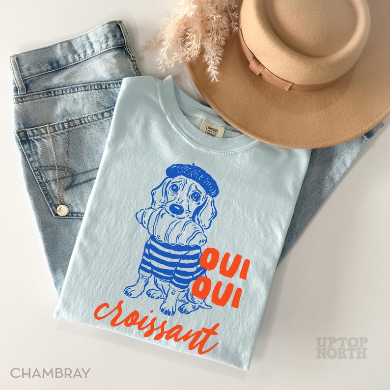 Peut inclure: Un t-shirt bleu clair avec un dessin d'un chien portant un b&eacute;ret et une chemise ray&eacute;e, tenant un croissant. Les mots "Oui Oui croissant" sont imprim&eacute;s en orange. Le t-shirt est associ&eacute; &agrave; un jean d&eacute;lav&eacute; et un chapeau beige.