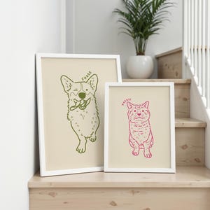May include: Two framed art prints featuring pet illustrations. One print shows a green outline of a dog with the name "Chase" above it. The other print shows a pink outline of a cat with the name "Maggie" above it. Both prints have a cream-colored background and white frames.