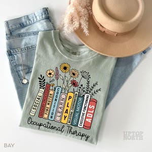 May include: A light green t-shirt with a graphic of a stack of books with the words "Occupational Therapy" and the names of different therapy areas on each book. The books are surrounded by flowers.