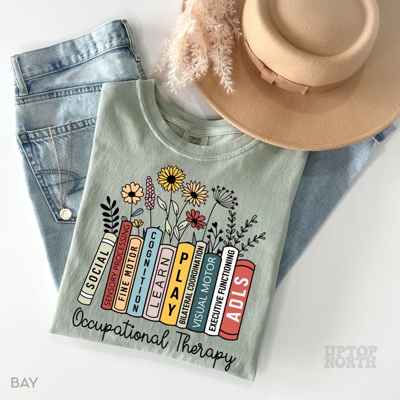 May include: A light green t-shirt with a graphic of a stack of books with the words "Occupational Therapy" and the names of different therapy areas on each book. The books are surrounded by flowers.