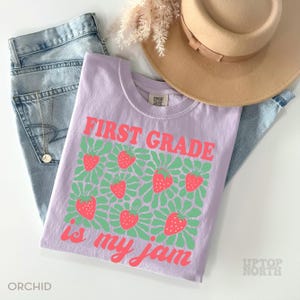 May include: A light purple t-shirt with the text "FIRST GRADE is my jam" in coral. The shirt features a pattern of strawberries and green floral designs. The shirt is paired with blue jeans and a tan hat.