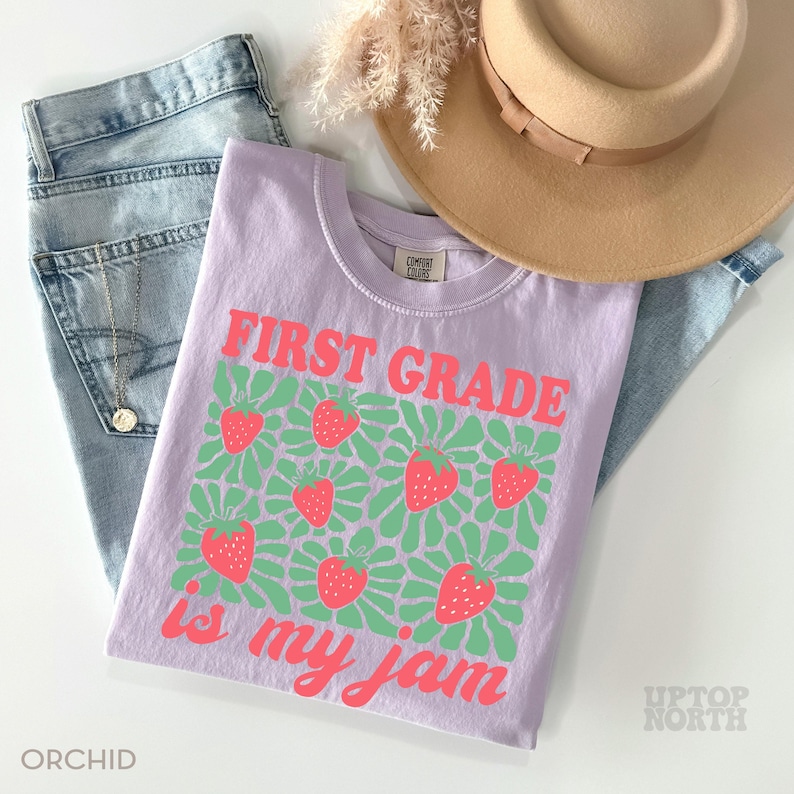 May include: A light purple t-shirt with the text "FIRST GRADE is my jam" in coral. The shirt features a pattern of strawberries and green floral designs. The shirt is paired with blue jeans and a tan hat.