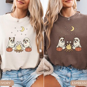May include: Ivory and espresso-colored t-shirts featuring a graphic of two dogs dressed as ghosts roasting marshmallows over a campfire, with pumpkins, a crescent moon, and stars. The shirts are made of soft material.