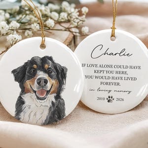 May include: Two round white ceramic ornaments with gold hanging strings. One features a colorful illustration of a Bernese Mountain Dog. The other displays the text: Charlie, IF LOVE ALONE COULD HAVE KEPT YOU HERE, YOU WOULD HAVE LIVED FOREVER, in loving memory, 2010, a paw print, and 2026.