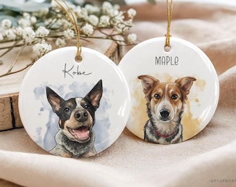 May include: Two white ceramic dog ornaments with gold string hangers. One features a black and white dog named Kobe, the other a brown and white dog named Maple. The ornaments are round and have watercolor-style portraits.