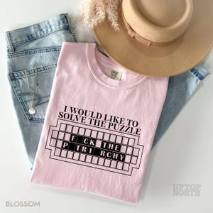 May include: Pink t-shirt with the text "I would like to solve the puzzle" and a black and white graphic of a crossword puzzle with the words "FCK THE TRCHY" in the grid.