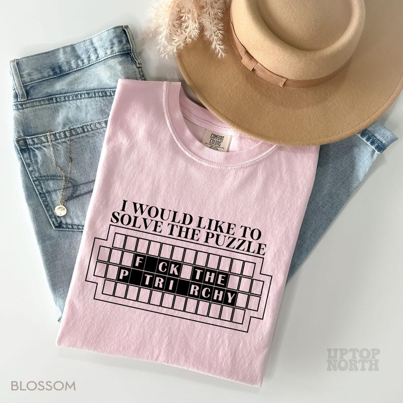 May include: Pink t-shirt with the text "I would like to solve the puzzle" and a black and white graphic of a crossword puzzle with the words "FCK THE TRCHY" in the grid.