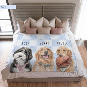May include: A light blue and white checkered throw blanket featuring illustrations of three dogs with the names NOVA, SUNNY, and ARCHIE printed above them. The blanket displays a black and white dog, a golden dog, and a golden dog holding a ball.