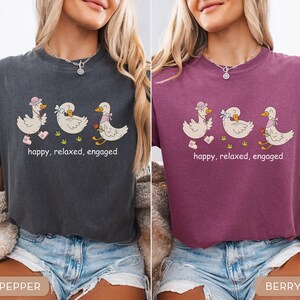 May include: Two t-shirts, one pepper and one berry, each featuring a cartoon design of three ducks. The ducks are wearing hats, bows, and scarves, and are walking on grass. The text "happy, relaxed, engaged" is below the ducks.