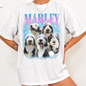 May include: A white short-sleeve t-shirt featuring a graphic print of five black and white dogs with bandanas. The text MARLEY is printed in a blue and pink gradient font above the dogs, set against a light blue cloudy background.